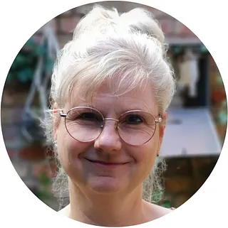 Lächelnde Frau mit grauen Haaren und runder Brille.