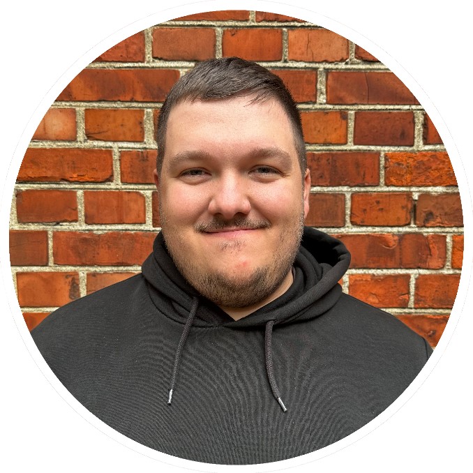 Lächelnder Mann mit kurzen braunen Haaren im dunklen Kapuzenpulliund Brille.