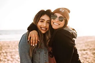 Zwei lächelnde Frauen umarmen sich am Strand.